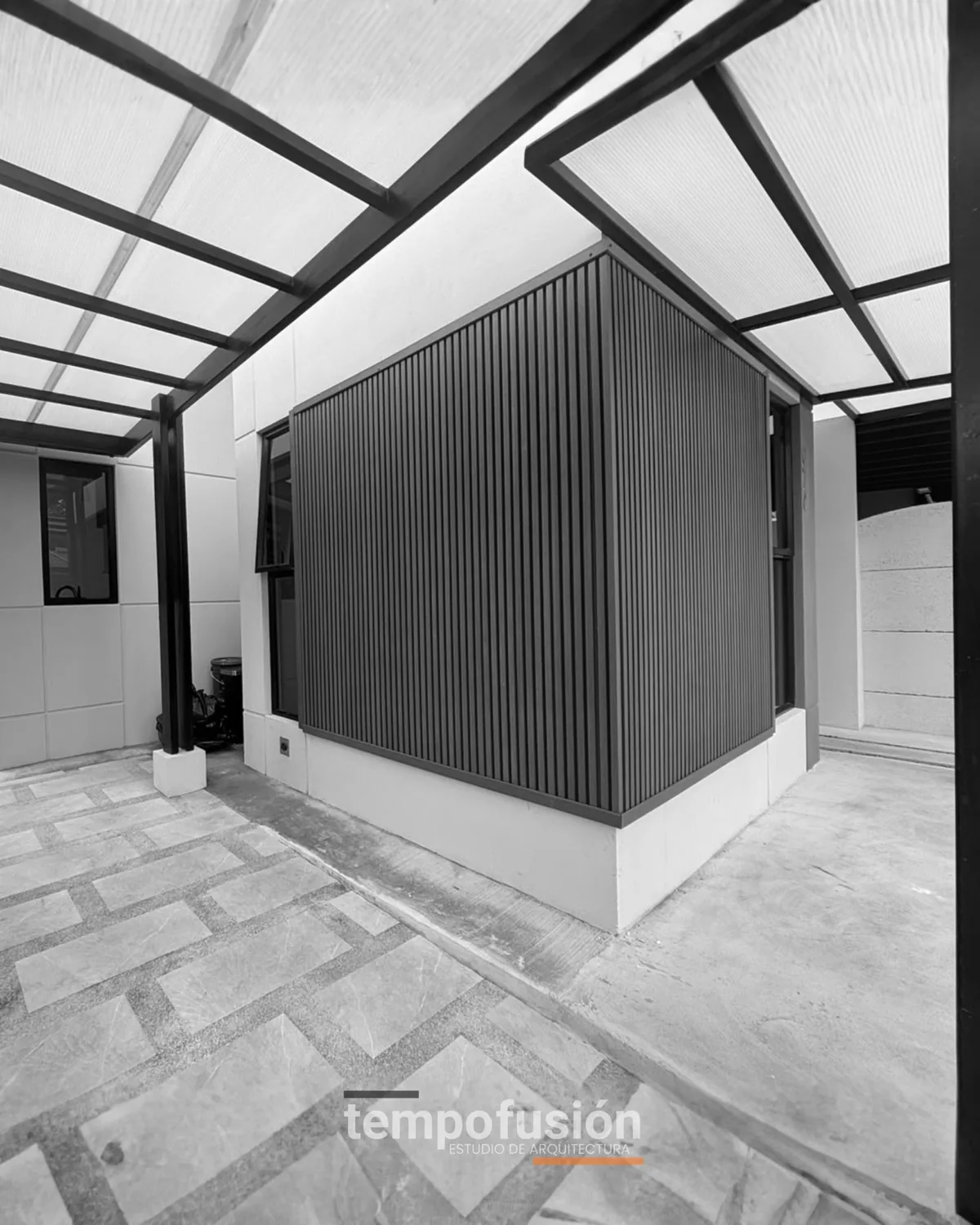 Pasillo exterior techado con cubierta traslúcida y celosía metálica, en blanco y negro